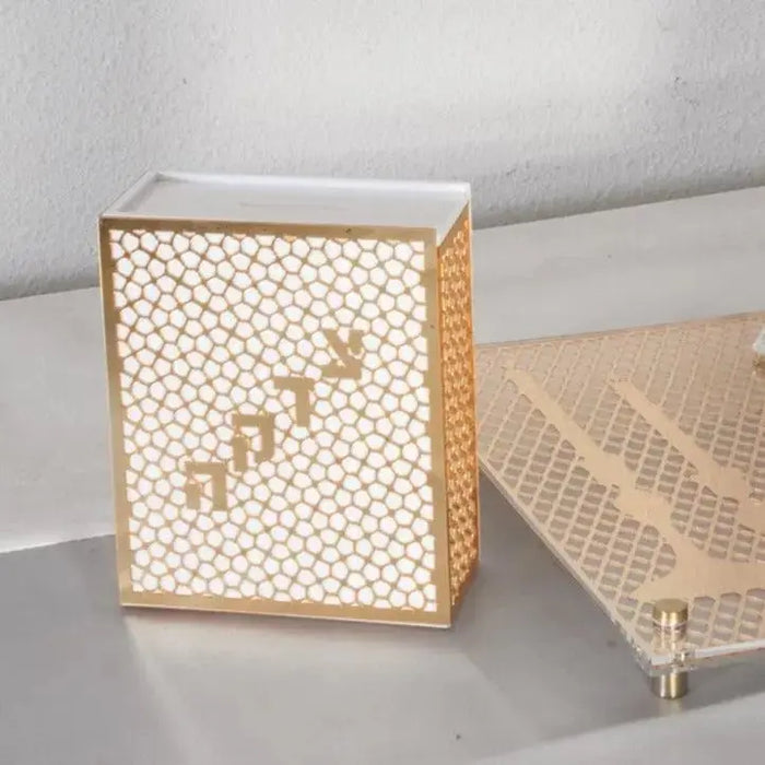 Laser cut tzedakah box with geometric pattern in natural wood and white, featuring Hebrew letters, displayed on table with matching woven placemat.