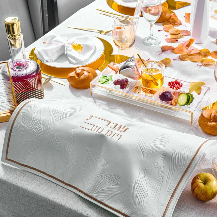 Waterdale Collection white leaf patterned challah cover with gold trim displayed on Shabbat table with place settings and fresh produce.
