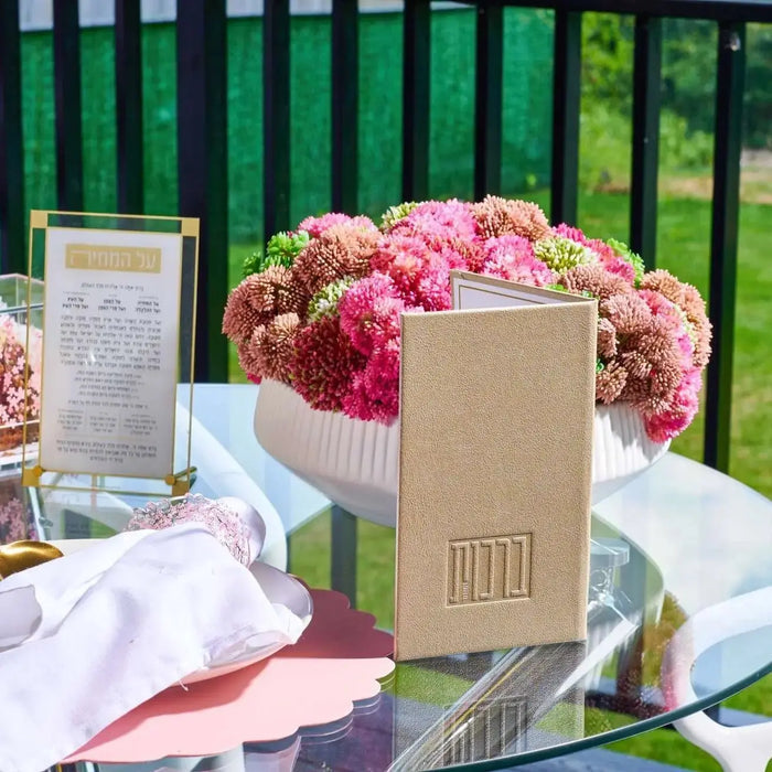 Tan leather Birchas Hashachar booklet displayed on outdoor table with pink flowers and white linens in background