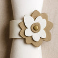Tan and cream leather flower napkin wrap with layered petal design and gold button center, displayed on white napkin.