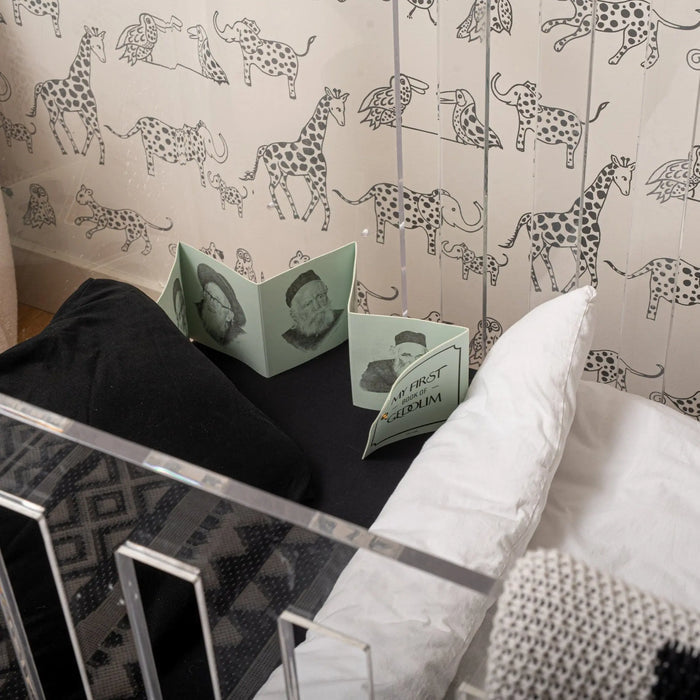Leather Kids Gedolim Book in black leatherette displayed on white bedding with patterned wallpaper background, showing prayer book cover design.