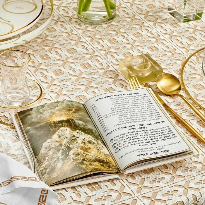 Leather Painted Haggadah with gold accents open on patterned table, displaying illustrated pages and text for Passover celebration
