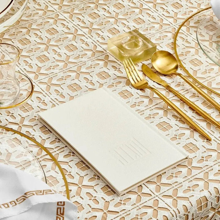 White leather Painted Haggadah displayed on patterned tablecloth with gold utensils and place settings
