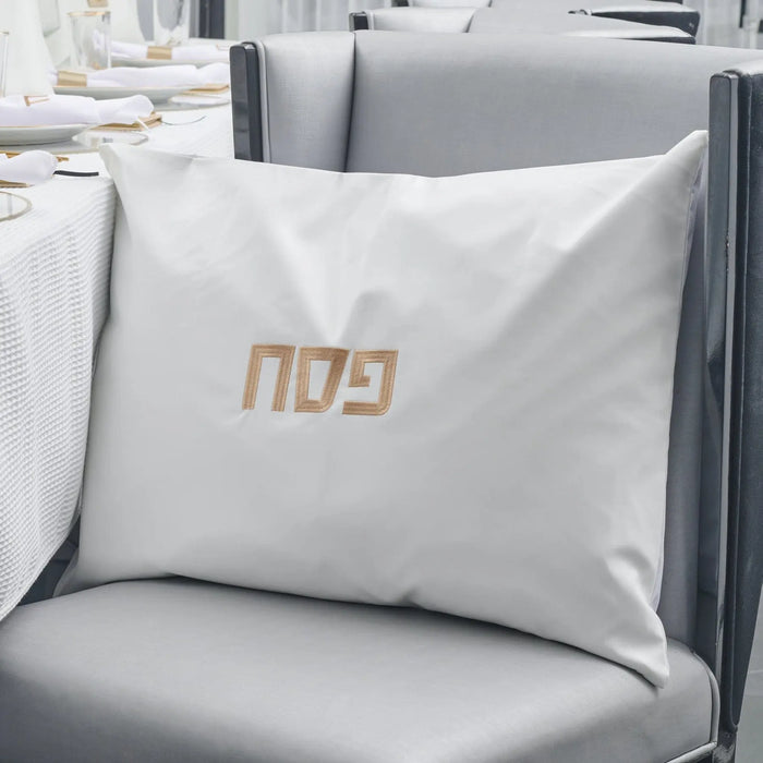 White leather pillow case with gold embroidered design, displayed on gray chair in modern interior setting