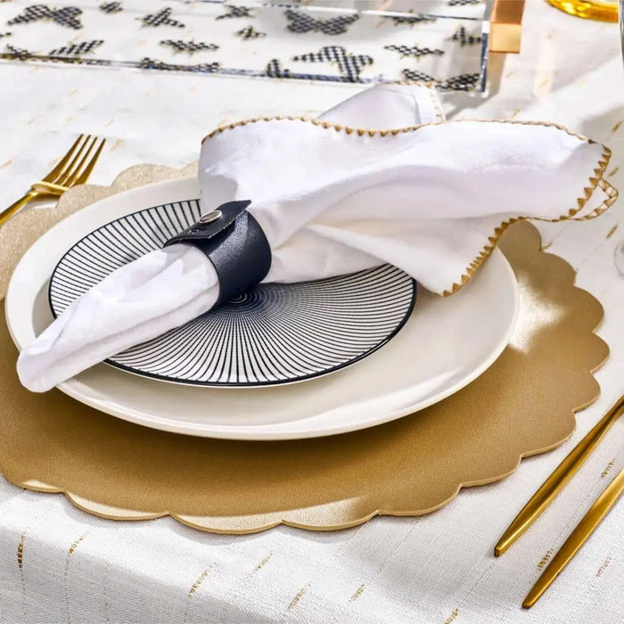 Black-bordered leather scalloped charger plate with silver accent rim, styled with gold flatware and napkin ring on white tablecloth.
