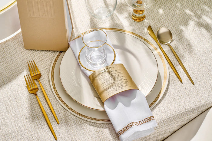 Gold leather Seder card napkin wraps arranged on white plate setting with place cards and gold flatware for Pesach table décor
