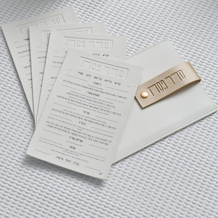 Cream leather Seder pouch with gold snap closure and printed Passover cards fanned beside it on white textured background.