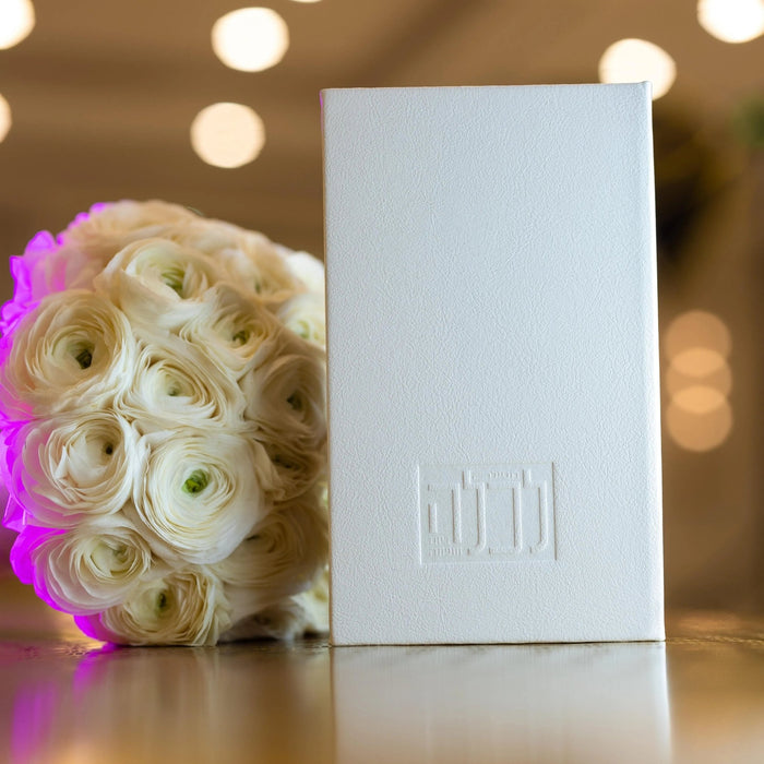 White leather Tefillah L'kallah booklet displayed next to pink and white roses with warm bokeh lighting