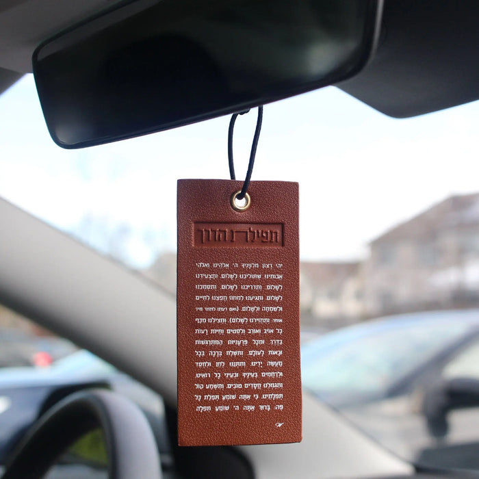 Brown leather Tefillas Haderech car hanger with Hebrew text, suspended from rearview mirror in vehicle interior.