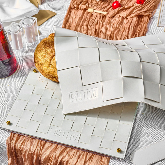 Silver leather woven challah cover with geometric square pattern design displayed on white surface with gold accents and tableware nearby.