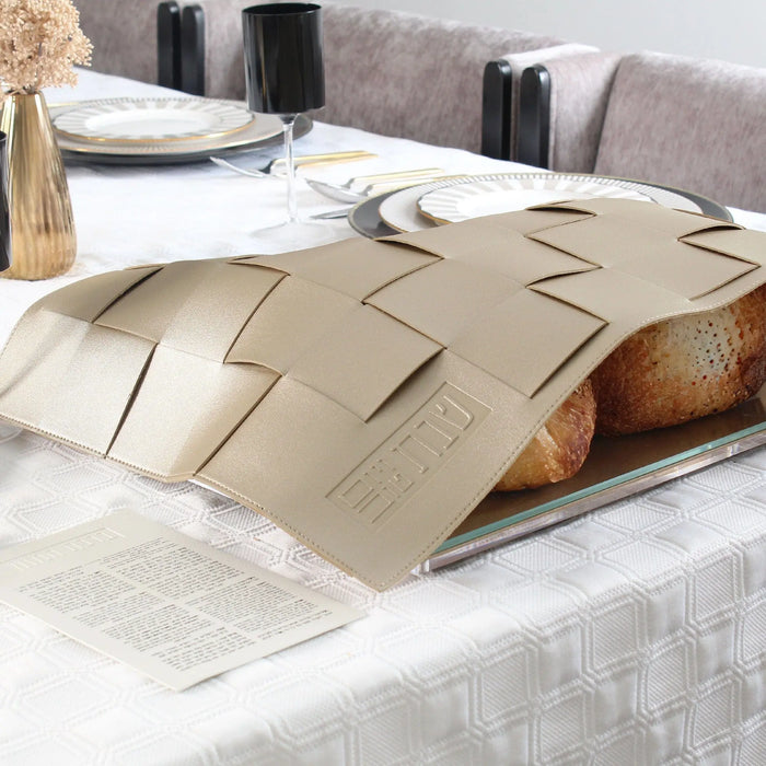 Tan woven leather challah cover with geometric cutout pattern, displayed on white bedding with place settings and wine bottle in background.