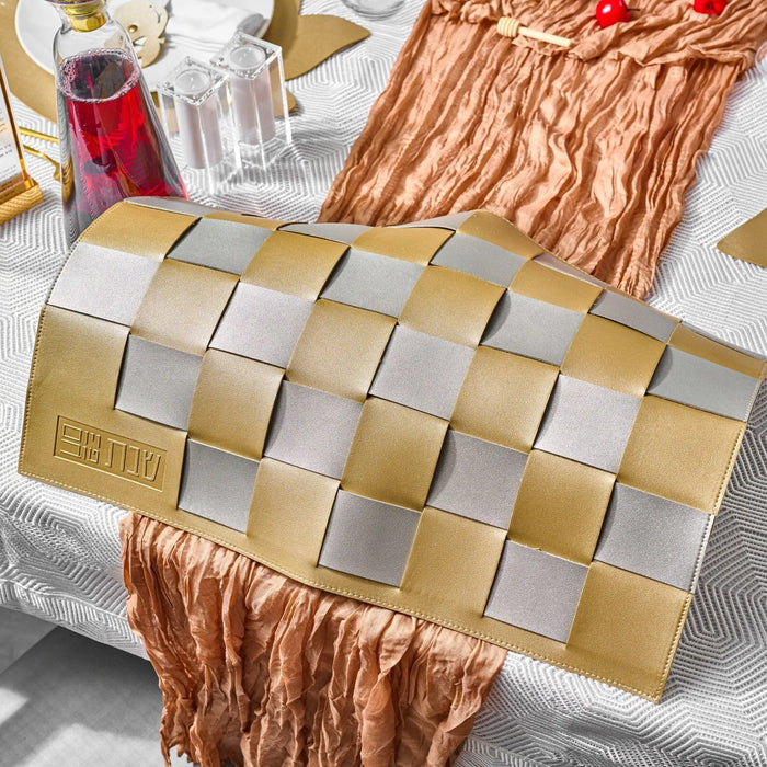 Tan leather woven challah cover displayed flat with copper-colored fabric accents, shown with coordinating tabletop items on a light surface.