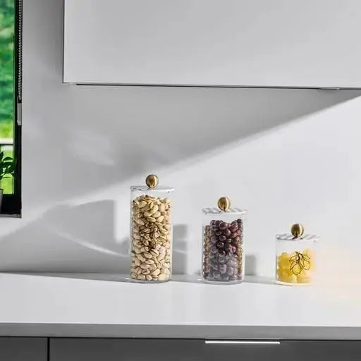 Three clear glass Lemona cylinder canisters in graduated sizes with brass lids, displayed on white shelf against geometric wall backdrop