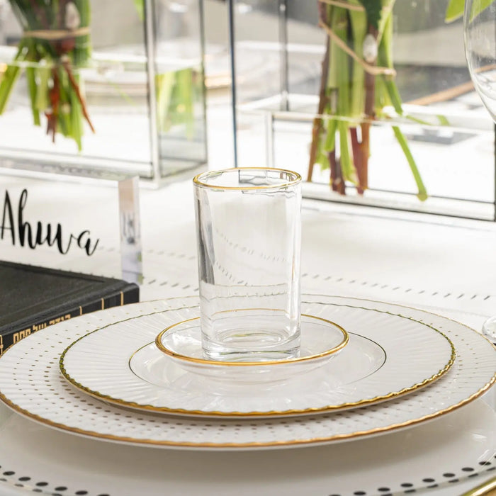 Modern glass kiddush cup with gold rim on layered white plate, set for Passover celebration with fresh greenery in background