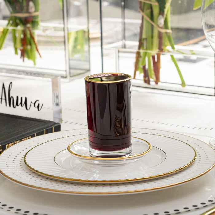 Modern glass kiddush cup filled with deep red wine on a decorative ceramic saucer with gold trim detailing, displayed on white tableware with greenery in background.