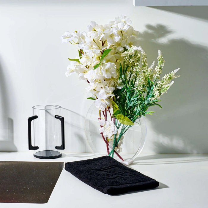 Modern white vase with black base holding white flowers and greenery on white tabletop surface.