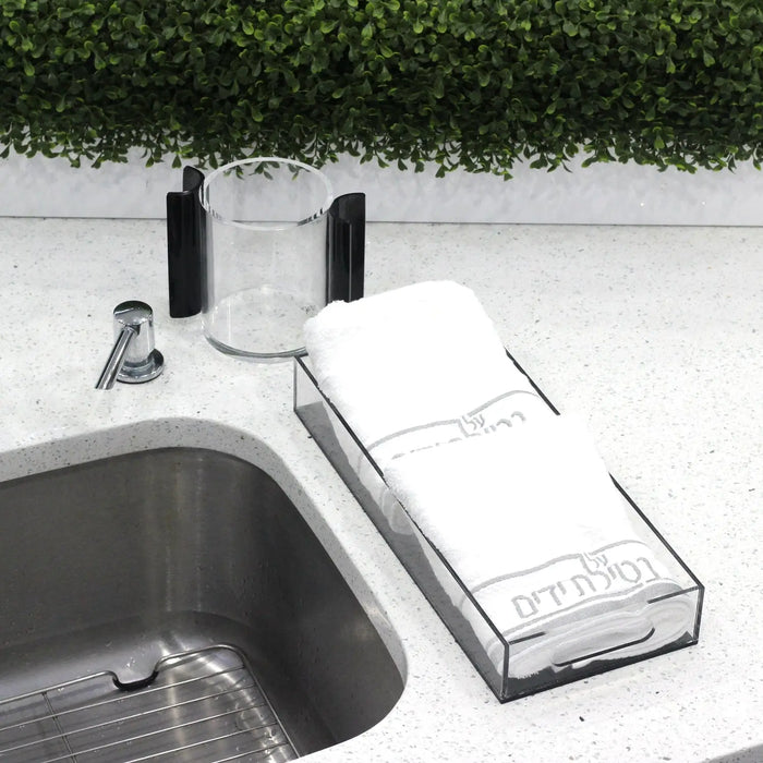 Black Netilas Yadayim hand towel displayed on white countertop near sink with soap dispensers, ideal for daily washing rituals.
