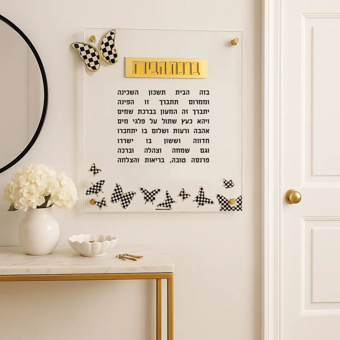 Onyx butterfly wall art displaying Birchas Habayis prayer text in black and gold, mounted above a modern shelf with home decor accessories.