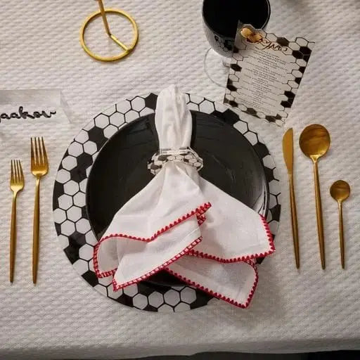 Black onyx charger plate with scalloped white beaded rim, gold cutlery, and red napkin on neutral textured surface.