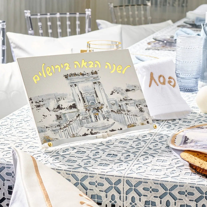 Painted Bais Hamikdash Haggadah stand displaying illustrated Jerusalem design, displayed on table with Passover Haggadah during Pesach seder meal setup.