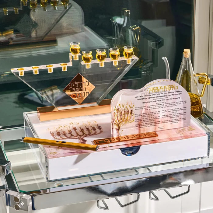 Hanukkah candle lighting set with a Lucite menorah, gold oil jug, a lighter and a printed brachos card on a glass shelf