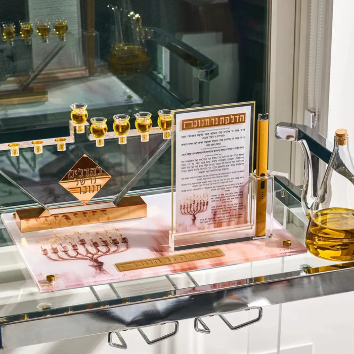 The Painted by Batya 2.0 Menorah Tray on a glass shelf with filled oil cups, an oil jug and a brachos plaque, set for Chanukah