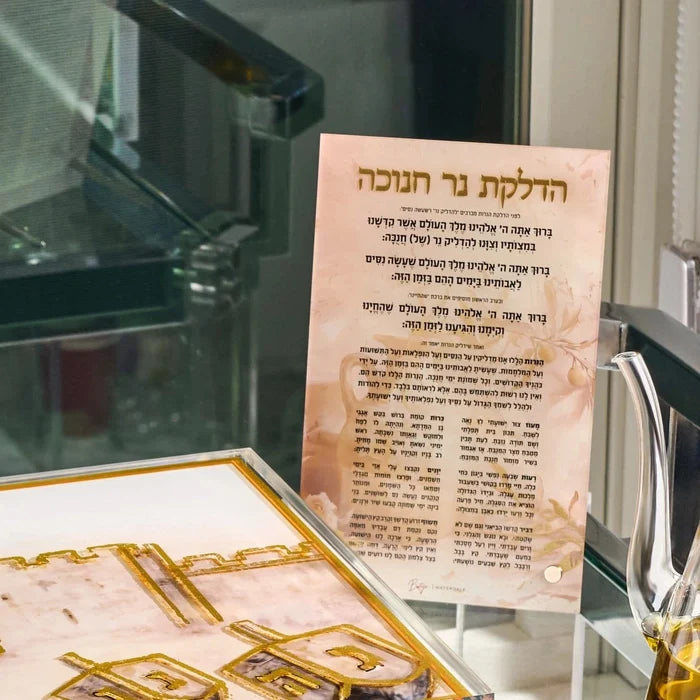 The Painted by Batya Chanukah brachos card standing in a clear holder, set out beside the menorah for Hanukkah candle lighting