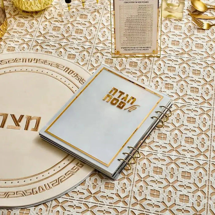 Painted by Batya lucite Haggadah with gold foil lettering displayed on decorative fabric with matching plate and bowl nearby.