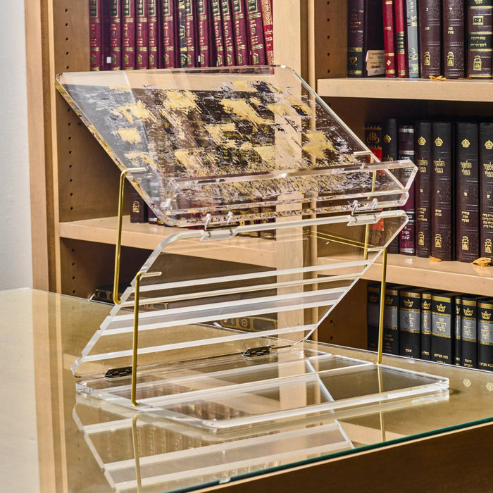 Clear acrylic foldable shtender with painted Jewish artwork displayed on a tabletop bookshelf