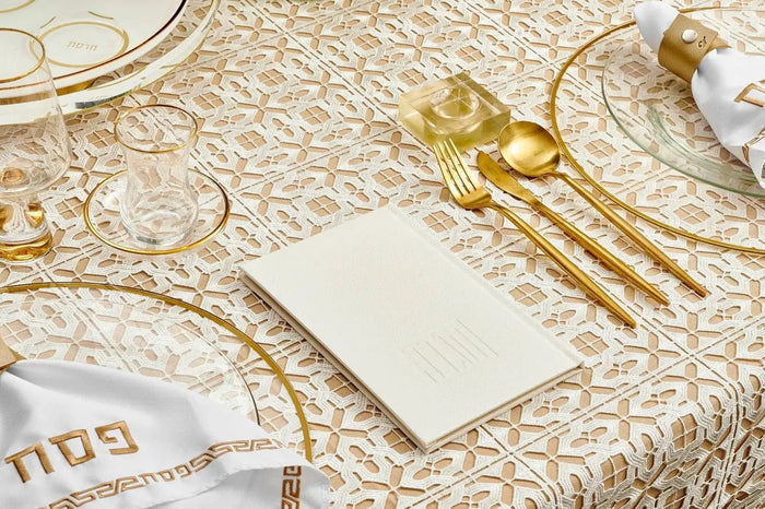 Painted Hardcover Leather & Lucite Haggadah Set in white and gold displayed with matching gold tableware on a textured cream tablecloth.