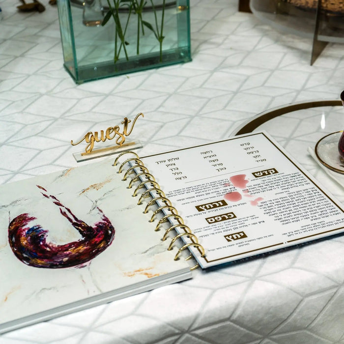 Painted Krias Yam Suf Haggadah displayed on marble surface with planner and gold accessories nearby