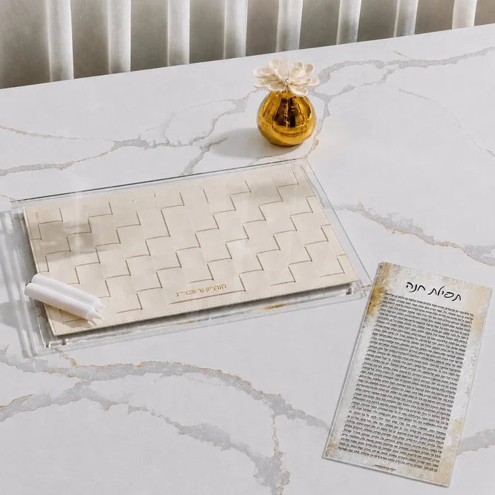A hadlokas neiros  tray set on a white marble table, featuring a clear acrylic base with a beige woven basket insert, two white candles, a small gold decorative vase, and a printed Hebrew tefilas chana card