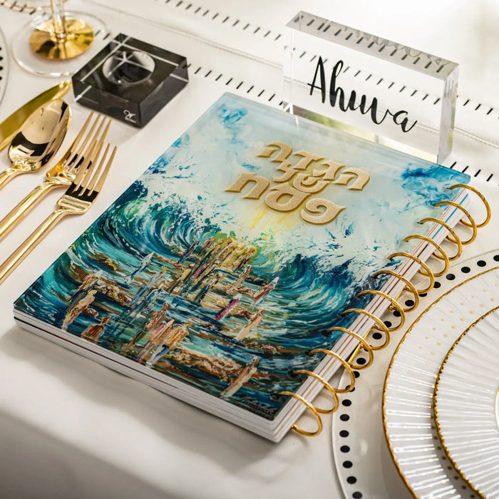 Painted Traditional Haggadah with blue watercolor landscape design, displayed alongside gold flatware and white dinnerware on a table setting for Pesach.