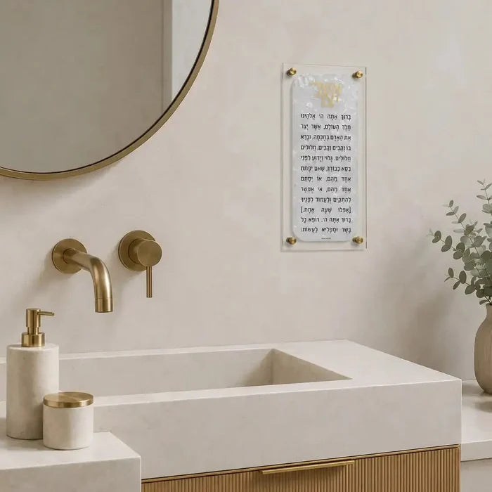 Pearlstone Asher Yatzar floating wall art in pearl marble finish displayed above a bathroom sink with brass fixtures and greenery.