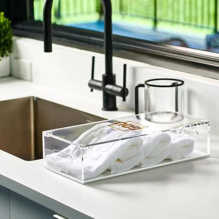 Clear lucite Pesach towel box with folded white towels and black faucet displayed on white kitchen counter near sink.
