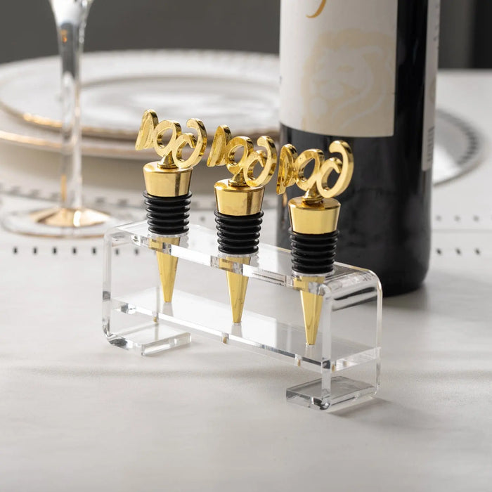 Four gold Pesach wine stoppers with clear lucite bases displayed in a row, paired with wine glasses and a bottle on a white surface.
