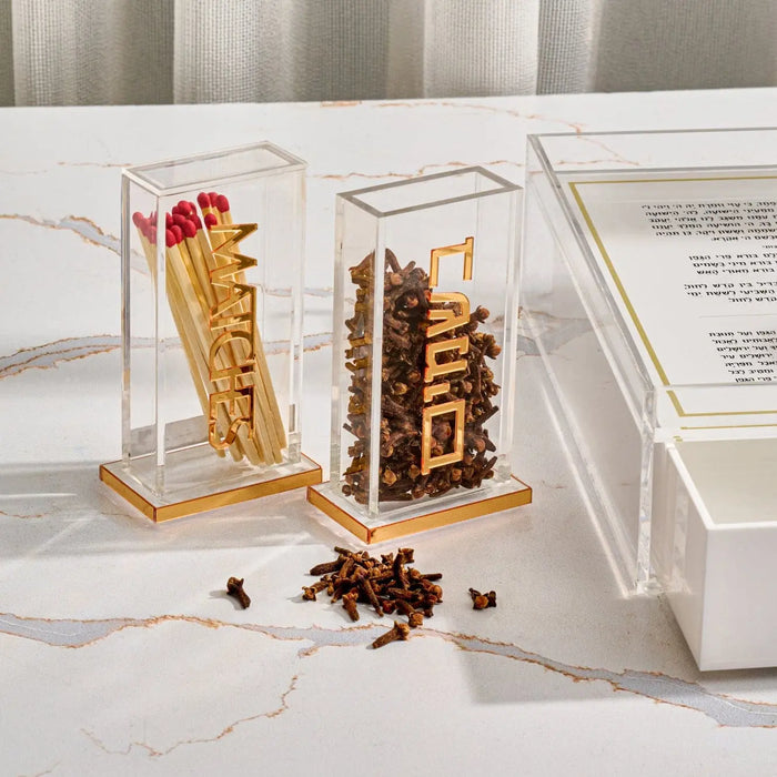 Two clear acrylic Pillar Besamim Holders displaying spice towers, with scattered cloves on marble surface and instructional cards nearby.