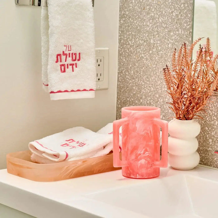 Pink resin Netilas Yadayim set displayed on wooden tray with towels and dried flowers in bathroom setting.