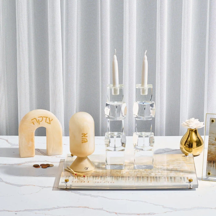 Cream resin arch tzedakah box with Hebrew text, displayed on clear acrylic base with candlesticks and gold vase for Shabbat.