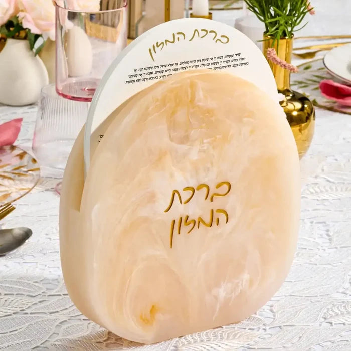 Cream-colored resin bencher set with Hebrew text, displayed on white surface with Shabbat tabletop styling and floral accents.