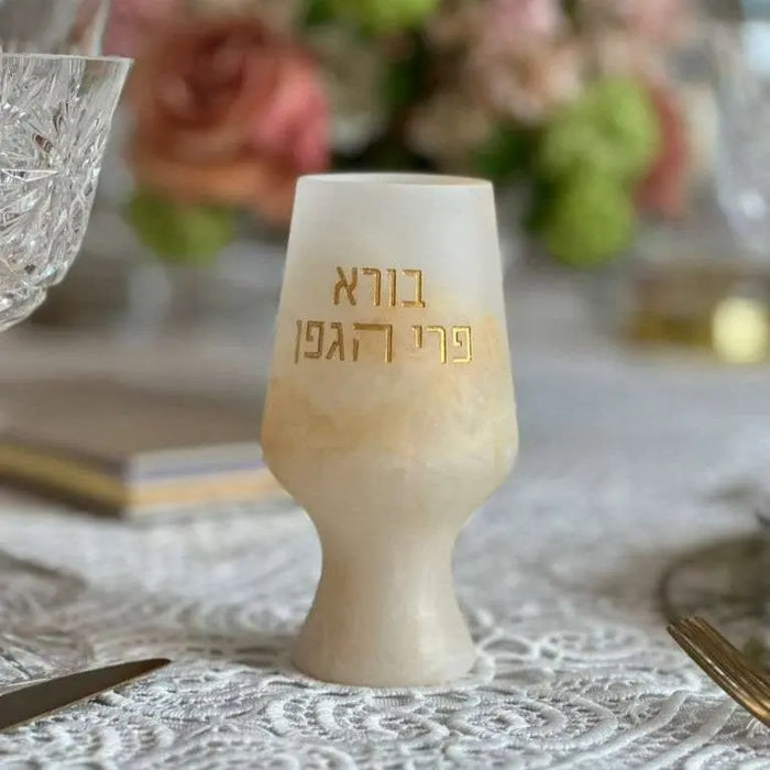 Cream-colored resin Kiddush cup with Hebrew text engraving, shown on a table setting with Havdalah ceremony items in soft focus background.