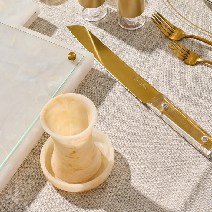 A close-up styled tabletop scene showing a gold-toned knife with a clear handle and a matching gold fork, alongside a gold resin  becher