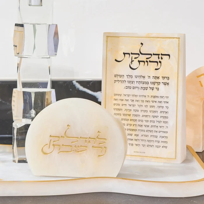 A small cream-colored  resin plaque with gold Hebrew lettering sits in front of a matching resin matchbo displayed on a white marble surface