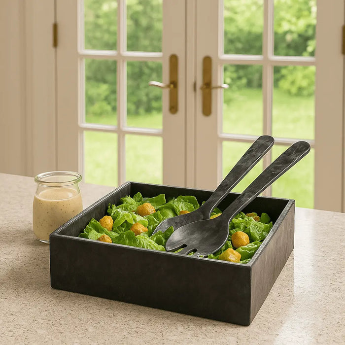 Black resin square salad bowl with gold accents and two matching servers displayed on a dining table near windows.