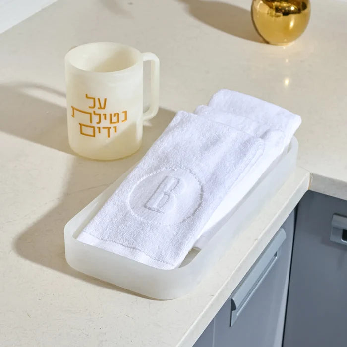 A white textured towel with an embroidered letter 'B' inside a circle, folded in a translucent white resin tray , set on a light beige countertop next to a cream-colored resin washing cup  with gold Hebrew text.
