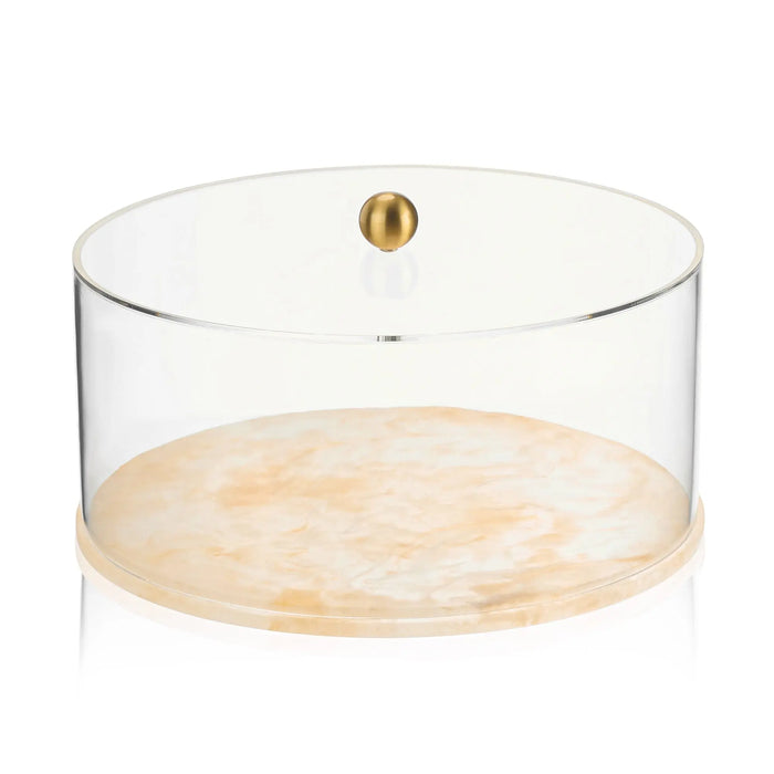 Round resin cake dome with clear acrylic cover and gold brass knob finial, featuring a marble or terrazzo-style base in cream and tan tones.