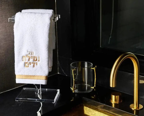Waterdale Collection S Towel Set displayed on black bathroom counter with gold faucet and glass, white towel with gold lettering