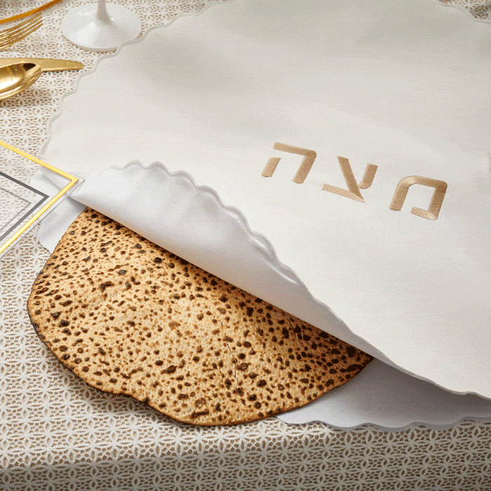 Scalloped edge Pesach set with white ceramic plate featuring Hebrew text and gold trim, displayed with matzah on neutral linen.
