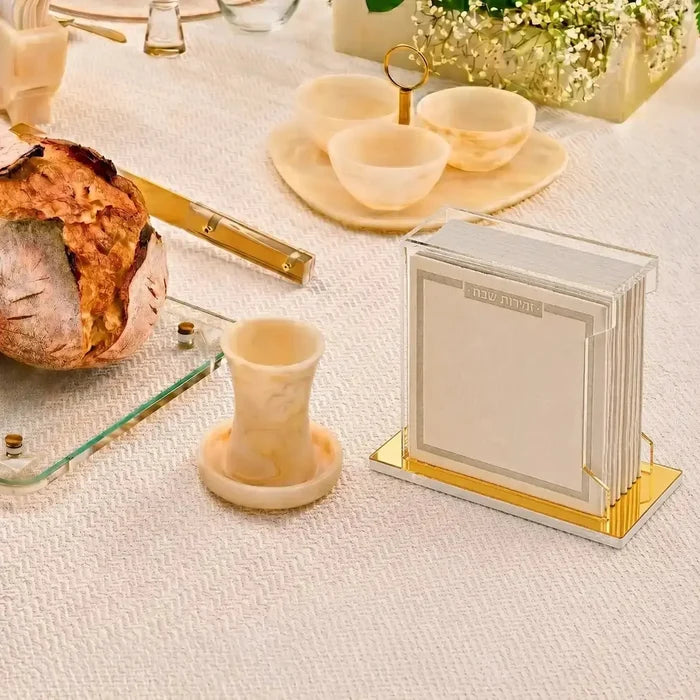 Cream and gold softcover leather bencher set displayed on white surface with Lucite stand, alongside challah bread and tableware for Shabbat.