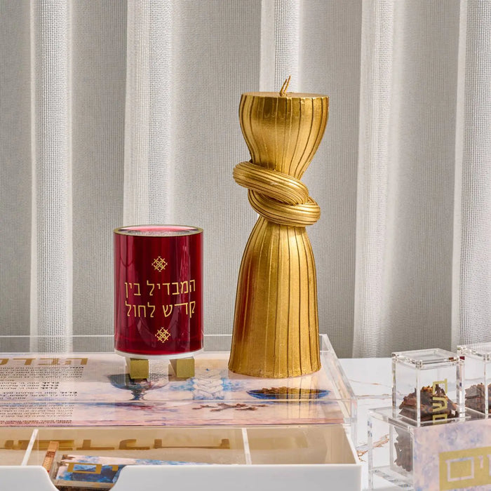 Spiral Havdalah candle in gold with twisted design, displayed next to a red Havdalah blessing candle on marble surface with white curtain backdrop.